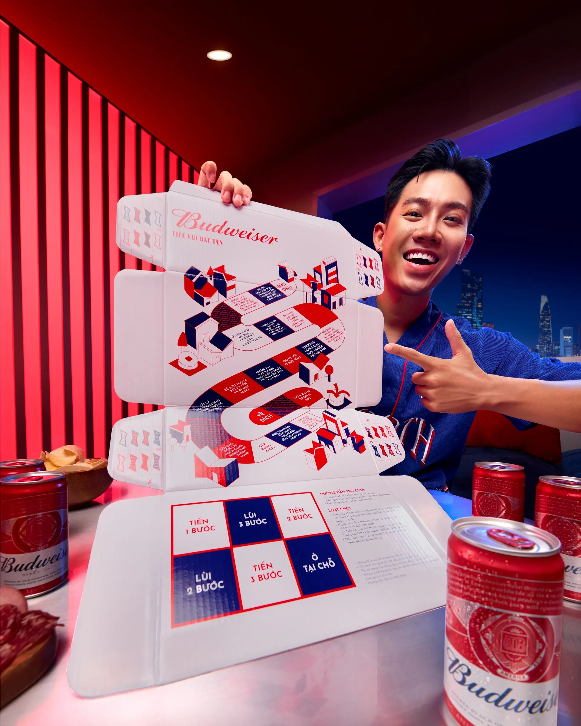 Một người đàn ông cười rạng rỡ bên các lon bia và hộp Budweiser với thiết kế đồ họa đỏ trắng.