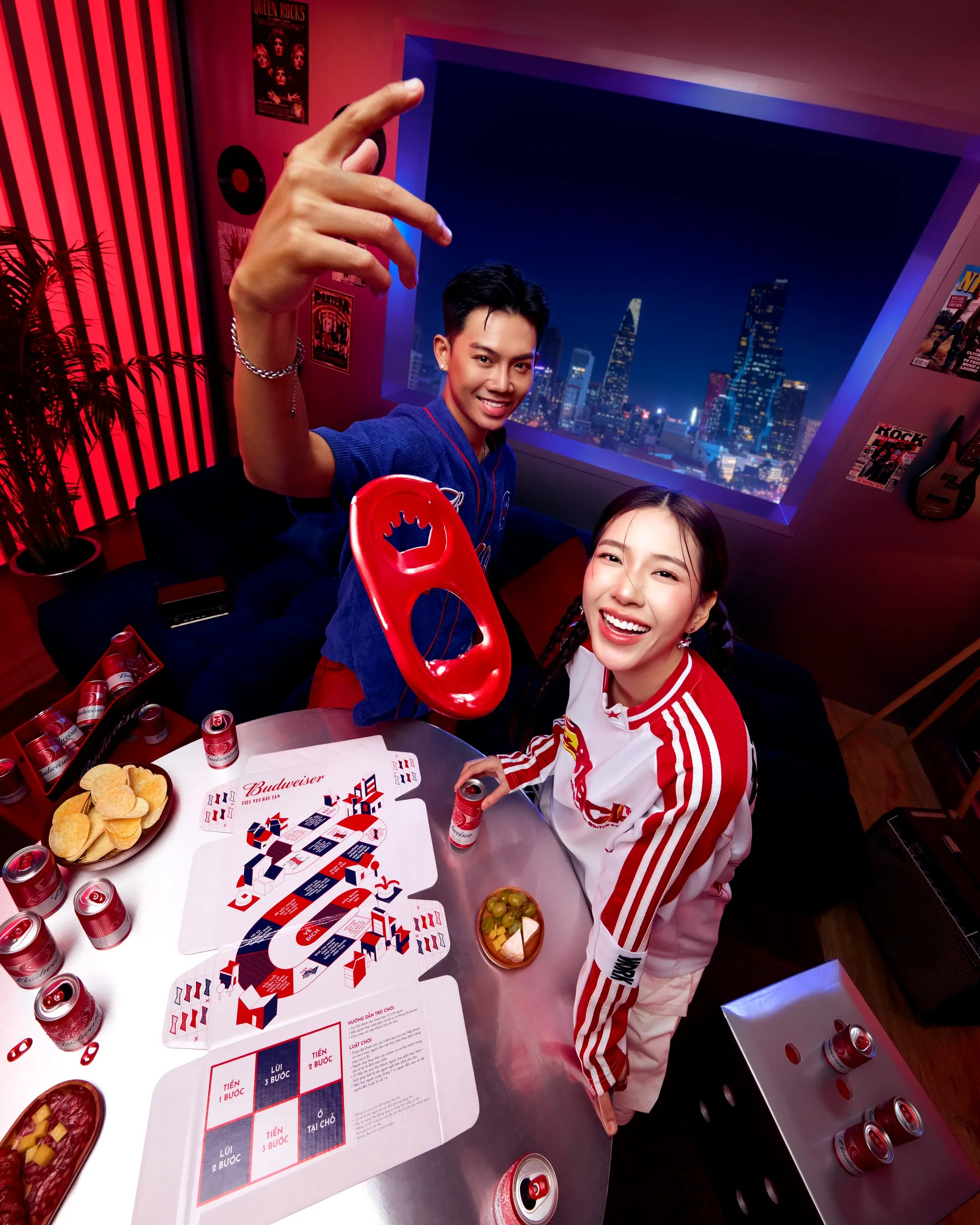 Nam và nữ chụp selfie trong phòng với đèn neon đỏ, trước màn hình thành phố đêm; trên bàn là bia Budweiser.