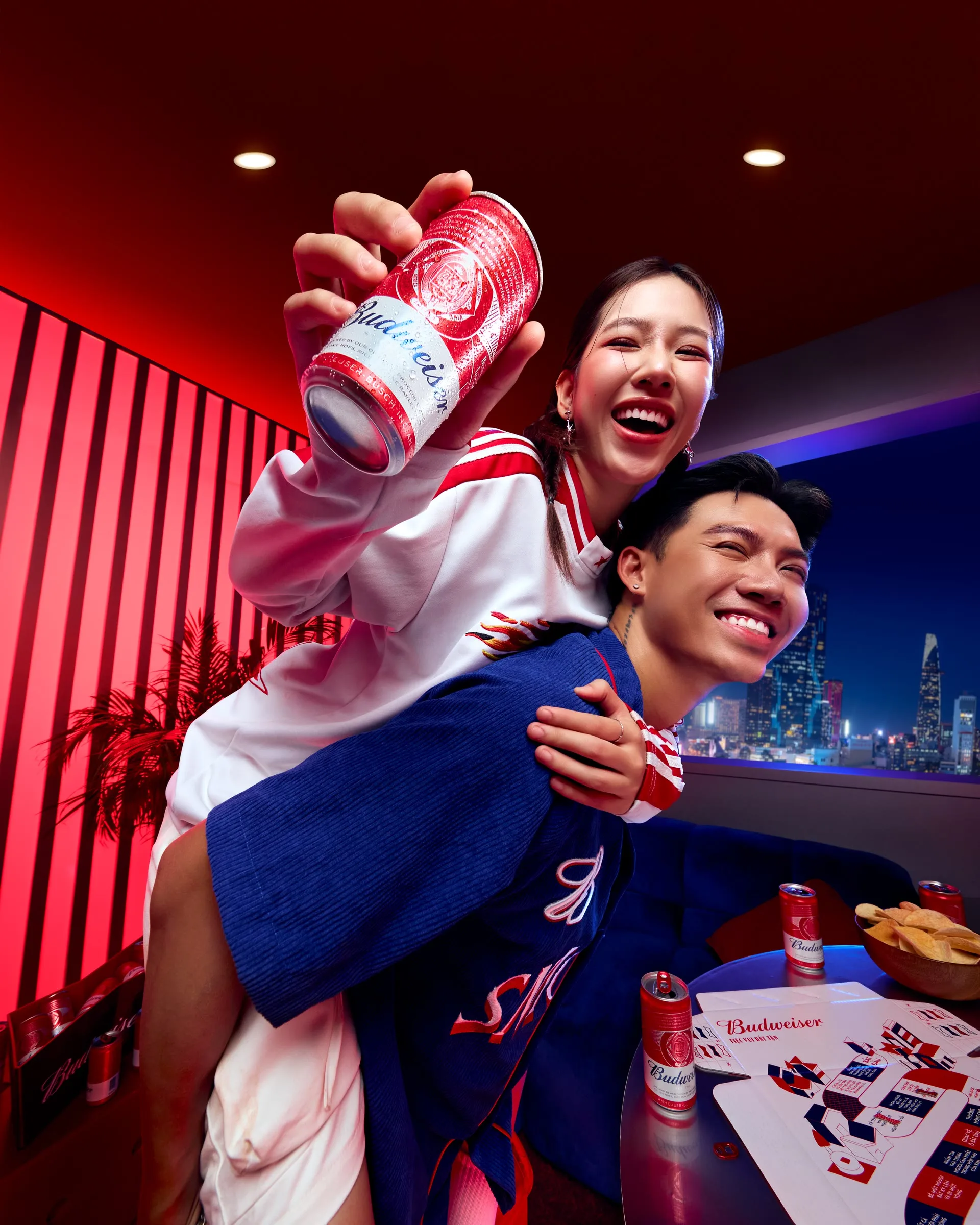 Hai người ôm nhau cười rạng rỡ với lon bia Budweiser trước cửa sổ nhìn ra thành phố về đêm.