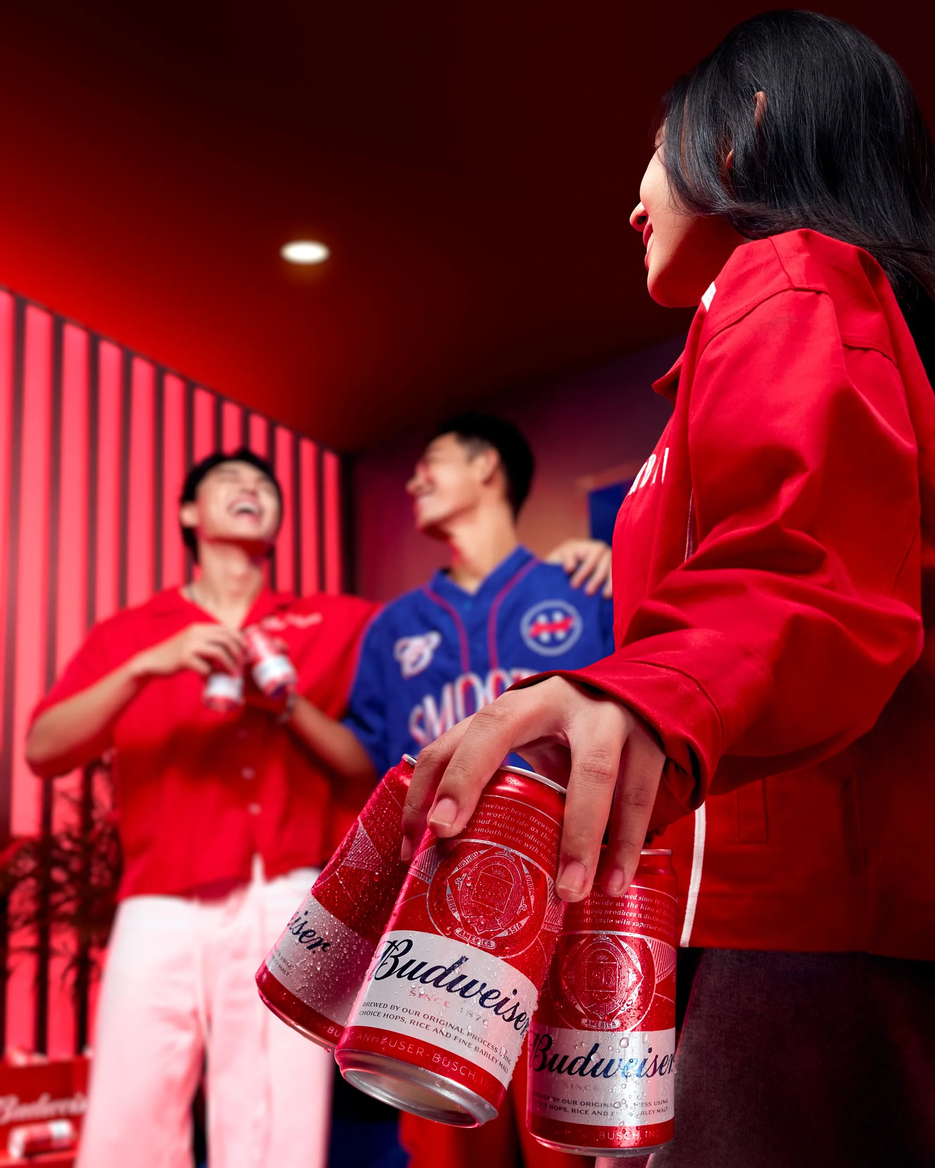 Cận cảnh một người phụ nữ cầm hai lon bia Budweiser trước khung hình rực đỏ với nhóm bạn đang vui vẻ ở phía sau.
