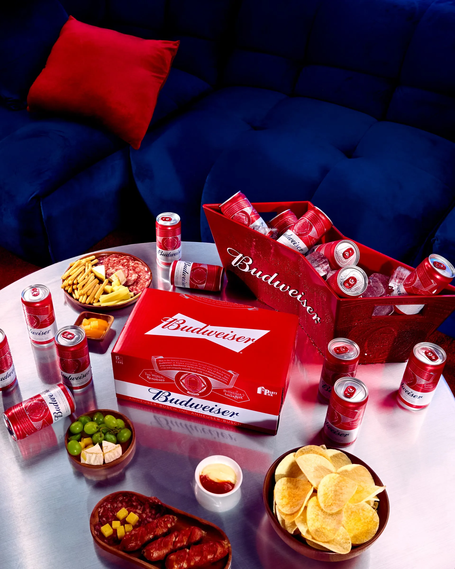Bàn tiệc với nhiều lon và hộp Budweiser đỏ trên nền ghế sofa xanh, kèm đĩa gà rán và khoai tây chiên.